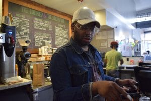 black man with a baseball cap and jean jacket putting coffee into the espresso machine